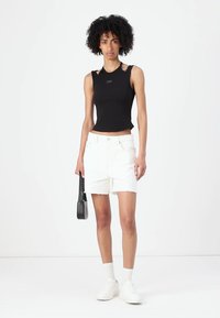 Black sleeveless top with a cropped design, paired with white denim shorts. Model wears white sneakers and carries a small black bag.