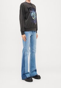 Persona che indossa un maglione grafico nero con la scritta "HERO", jeans svasati blu bicolore e scarpe platform nere, in piedi contro uno sfondo bianco.