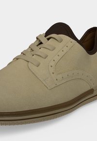 Chaussure beige en toile à lacets, avec détails perforés, col marron et semelle en caoutchouc texturé, vue de près de côté.