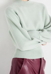 Personne portant un pull côtelé vert clair et un pantalon en cuir bordeaux taille haute, les bras croisés.