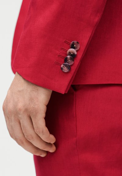 Κοντινό πλάνο του χεριού ενός ατόμου που ακουμπάει δίπλα σε ένα κόκκινο μανίκι σακακιού με τέσσερα σκούρα μαρμάρινα κουμπιά και ταιριαστό κόκκινο παντελόνι.