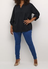 Chemisier noir à boutons, à manches longues, avec une coupe décontractée et des poignets retroussés, associé à un jean skinny bleu et des escarpins beiges.