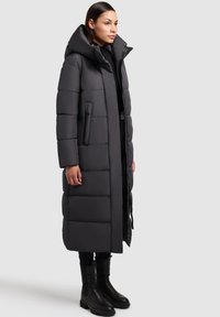 Długa, ciemnoszara kurtka puchowa z kapturem, z pikowanymi sekcjami, bocznymi kieszeniami oraz zamkiem z przodu, zestawiona z czarnymi botkami do kostki.