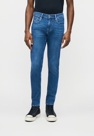 Muž oblečený v úzkych modrých rifliach a čiernych vysokých teniskách stojaci pred jednoduchým svetlým pozadím.