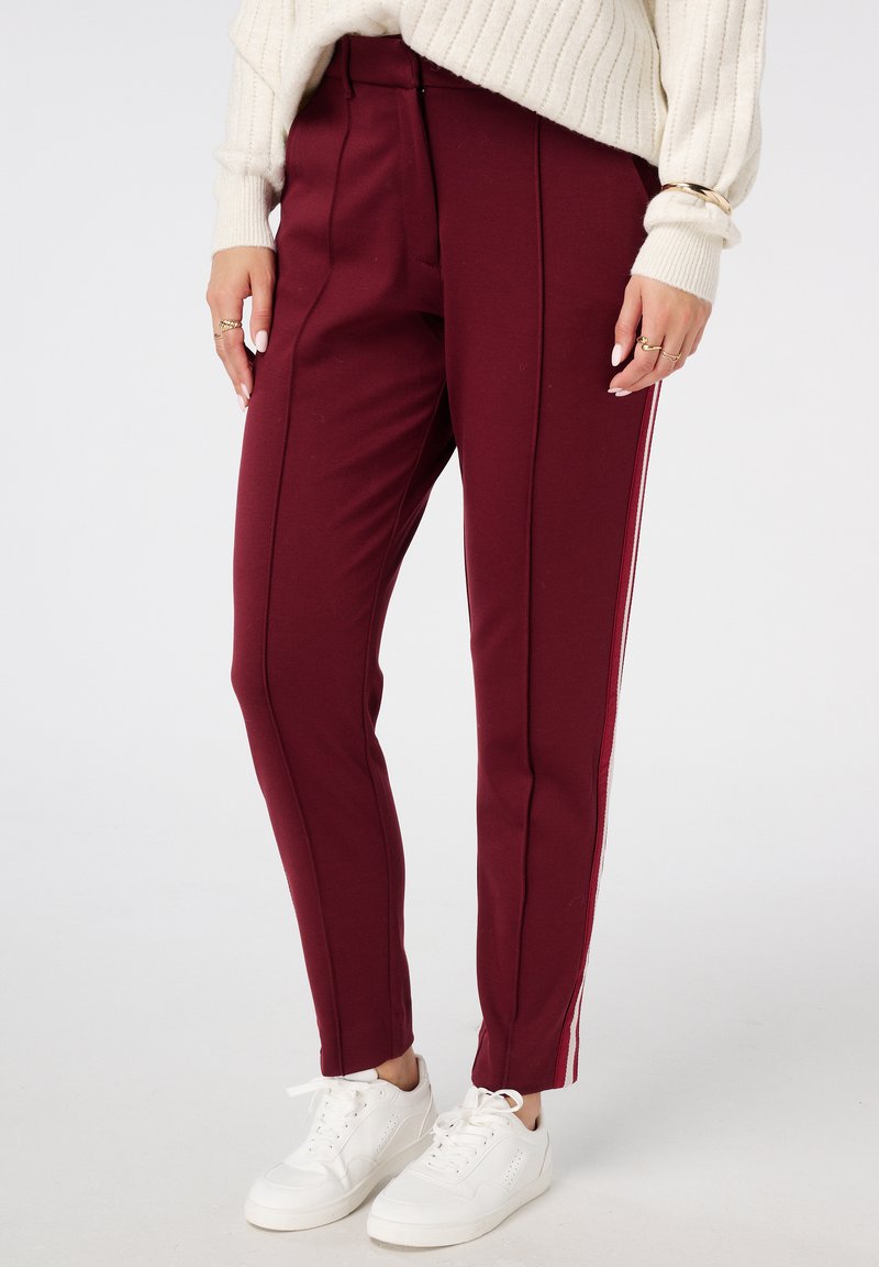 Pantalons bordeaux avec des coutures verticales, rayures latérales en blanc et rouge, fabriqués dans un matériau extensible, associés à des baskets blanches.