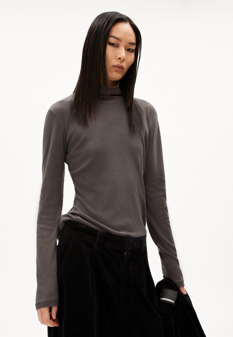 Junge Frau mit langen, glatten Haaren, die einen dunkelgrauen Rollkragenpullover und schwarze Hosen trägt und eine schwarze Jacke in ihrer rechten Hand hält.