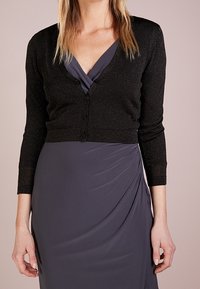 Svart, glittrande cardigan med långa ärmar och knappstängning, bärs över en draperad grå klänning med djup V-ringning.