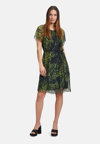 Durchsichtiges kurzes Kleid mit einem grün-schwarzen Zebra-Muster, tailliert mit einem Bindegürtel und mit schwingenden Ärmeln. Getragen mit schwarzen Plateauschuhen.