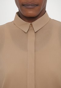 Beige button-up shirt with a smooth, slightly sheer texture, featuring a classic collar and a single button closure at the neck.