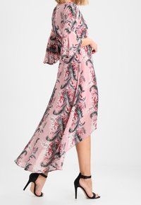 Różowa kwiatowa sukienka maxi z rękawami dzwonowymi, z czarnymi i czerwonymi wzorami liści. Noszona z czarnymi sandałami na wysokim obcasie. Asymetryczny dół.