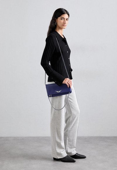 Blue leather crossbody bag with a chain strap, featuring a decorative wing accent. Worn with black ribbed button-up shirt and gray sweatpants.