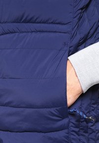 Námorně modrá péřová bunda s horizontálním prošíváním, vybavená boční kapsou a nastavitelnou šňůrkou. Měkký, lehký materiál.