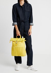 Mochila de malla amarilla con diseño de tapa enrollable, que cuenta con un cierre de cremallera y un bolsillo frontal. Llevada por una persona en un mono de mezclilla oscuro.