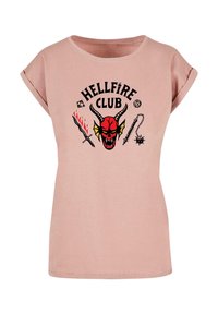 Camiseta de algodón rosa con mangas enrolladas, que presenta un diseño gráfico en negro y rojo de la cabeza de un demonio y armas, con el texto "HELLFIRE CLUB."