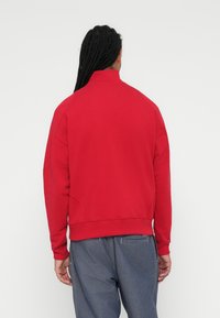 Sudadera roja con un alto cuello y puños acanalados. La tela parece suave con una textura lisa, combinada con pantalones de mezclilla holgados.