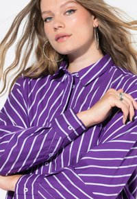Camicia a maniche lunghe violacea con strisce verticali bianche. Presenta un colletto e polsini con bottoni, realizzata in un tessuto liscio.