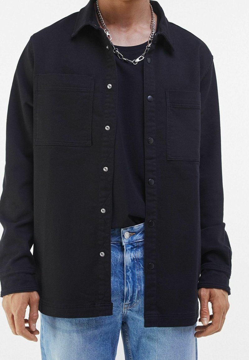 Zwart overhemdjack met twee borstzakken, knoopsluiting en metalen drukknoopsluitingen. Draagt over een zwart T-shirt en gecombineerd met lichtblauwe jeans.