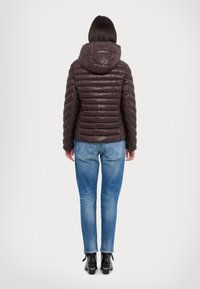 Doudoune marron foncé avec texture matelassée et capuche, portée sur un jean bleu et des bottines noires, vue de dos.