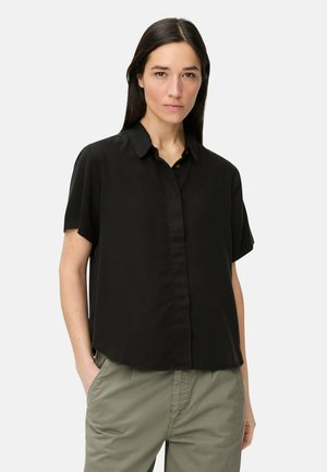 Kvinde med glat mørkt hår iført en løs sort kortærmet skjorte med knapper og olivengrønne bukser, står med hænderne i lommerne.