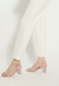 Sandales à talons bloc en paillettes rose doré avec un design à double bride et une boucle à la cheville, associées à un pantalon blanc ajusté.