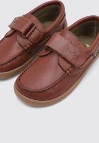 Zapatos de cuero marrón con una correa de velcro, que presentan costuras decorativas y una suela de goma. El diseño es plano con puntas redondeadas.