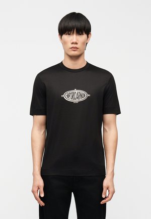 Jeune homme aux cheveux noirs lisses portant un t-shirt noir Emporio Armani avec un logo blanc centré sur la poitrine, debout devant un fond uni.