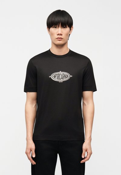 Jeune homme aux cheveux noirs lisses portant un t-shirt noir Emporio Armani avec un logo blanc centré sur la poitrine, debout devant un fond uni.