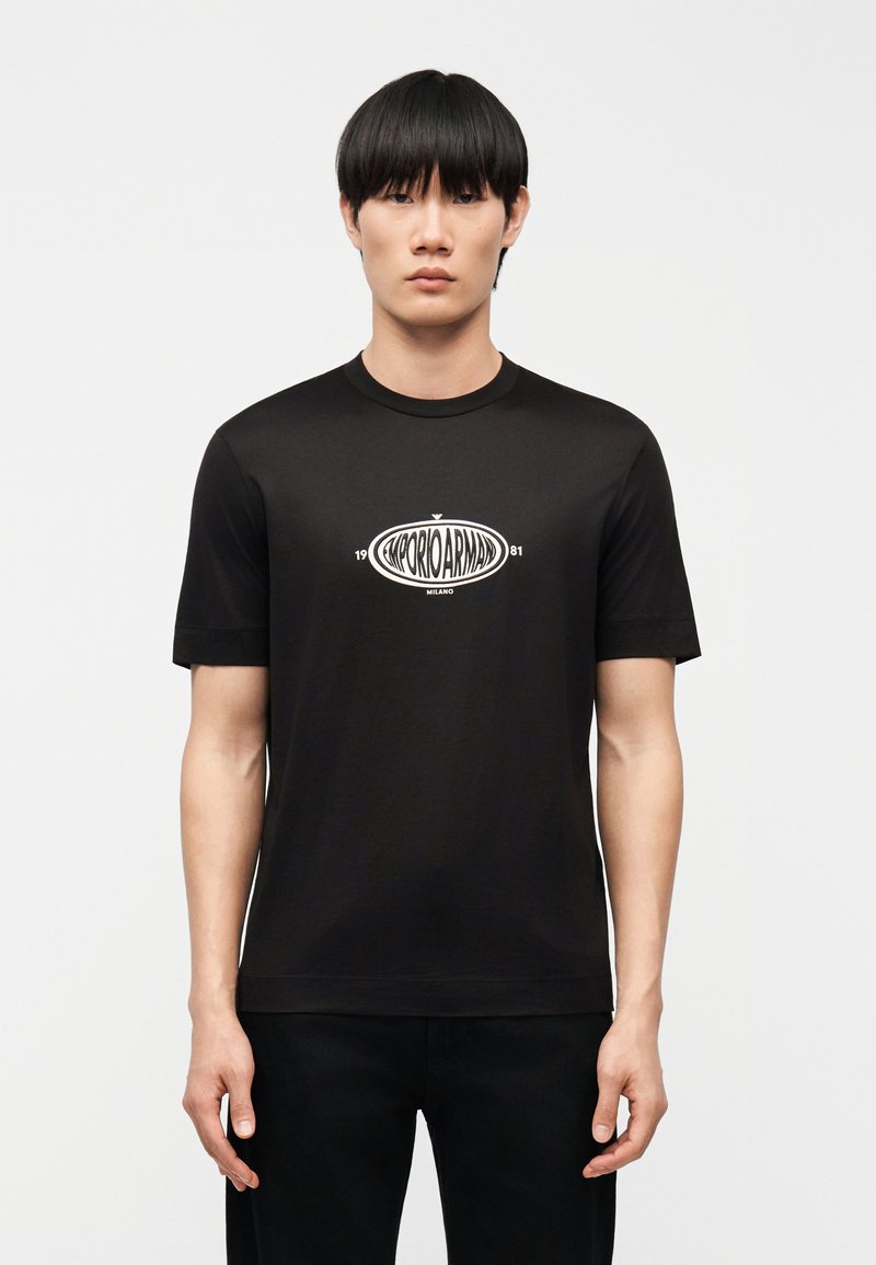 Νέος άνδρας με ίσια μαύρα μαλλιά που φοράει μαύρο t-shirt Emporio Armani με λευκό λογότυπο στο κέντρο του στήθους, στέκεται μπροστά σε απλό φόντο.