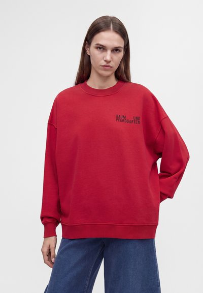 Femme aux cheveux bruns et raides portant un sweat-shirt rouge oversize avec un texte noir sur la poitrine et un jean bleu ample, debout devant un fond uni.