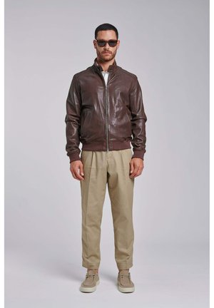 Uomo con giacca in pelle marrone scuro, pantaloni beige, occhiali da sole e scarpe beige, in piedi su uno sfondo grigio uniforme.