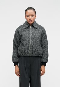 Veste bomber à motif pied-de-coq noir et blanc avec une fermeture éclair à l'avant, un large col et des poignets côtes, associée à un pantalon taille haute gris.