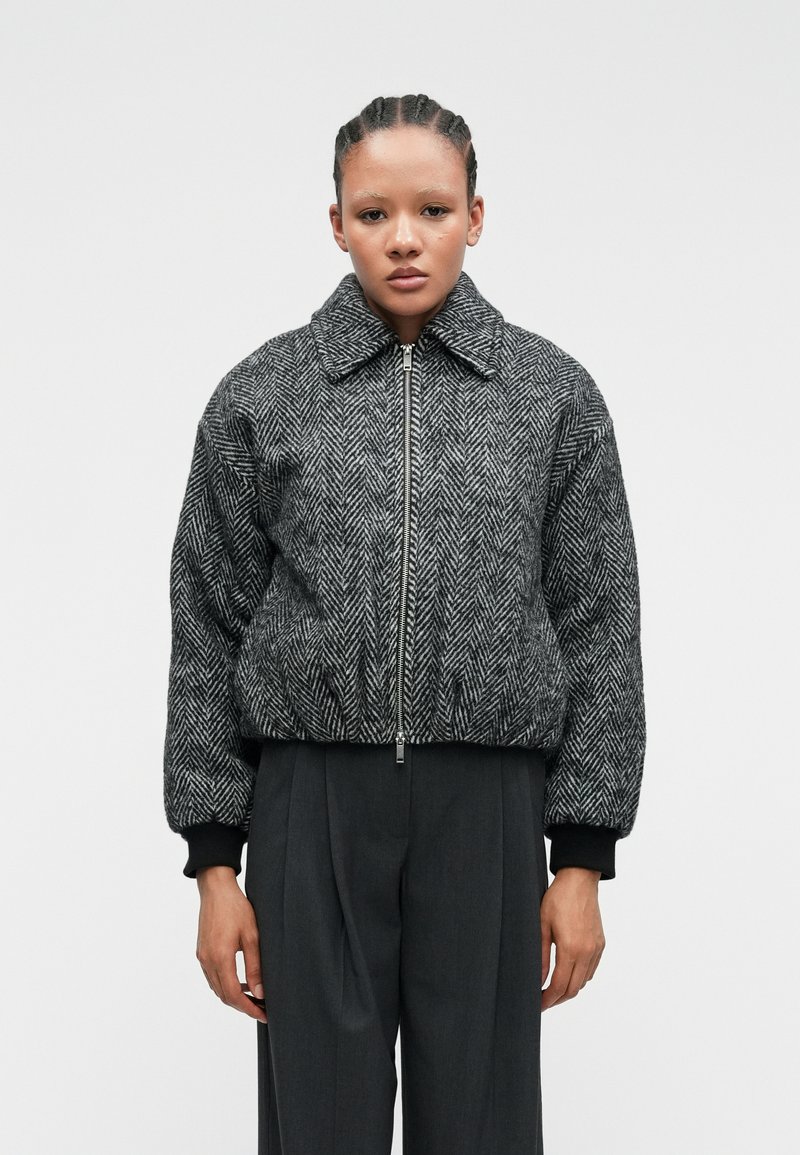 Veste bomber à motif pied-de-coq noir et blanc avec une fermeture éclair à l'avant, un large col et des poignets côtes, associée à un pantalon taille haute gris.