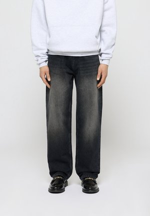 Person iført en lysegrå hættetrøje, løstsiddende falmede sorte jeans og sorte loafers med guldkædedetalje, stående mod en ensfarvet baggrund.