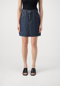 Falda de mezclilla en azul oscuro, con cintura alta, bolsillos frontales y corte recto. Llevada con mule negro de punta abierta.