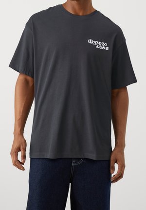 Czarny bawełniany t-shirt w ciemnoszarym kolorze, o luźnym kroju i oversize'owym fasonie, z białym nadrukiem na lewej piersi. Krótkie rękawy i okrągły dekolt.