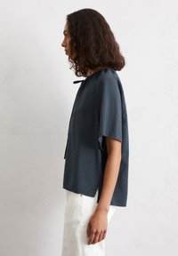 Top corto a maniche corte di colore grigio scuro con finitura testurizzata, scollatura rotonda e spacchi laterali. Indossato con pantaloni bianchi, mostrato di profilo.