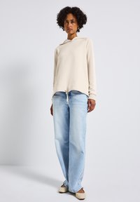 Felpa beige con cappuccio e cordino sull'orlo, abbinata a jeans azzurri larghi e scarpe basse color beige.