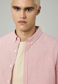 Chemise boutonnée rayée rouge et blanche avec un col étalé, portée avec un pull léger écru en dessous. Boutons plats et tissu texturé.