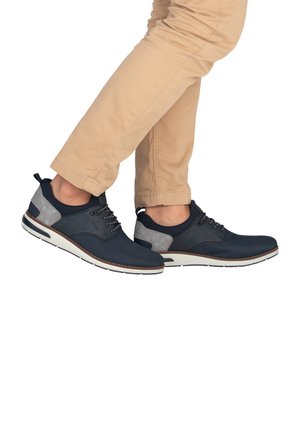 Mann in beigen Hosen und navyblauen Freizeitschuhen mit grauen Akzenten, der mit einem Bein über das andere steht.