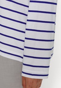 Primo piano di un braccio di una persona che indossa una camicia bianca a maniche lunghe con righe orizzontali blu equidistanti e pantaloni grigio chiaro.