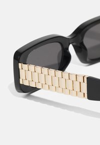 Schwarze Sonnenbrille mit rechteckigen Gläsern, ausgestattet mit einem goldenen Arm im Kettendesign mit strukturierten Oberflächen, die das elegante Rahmendesign ergänzen.