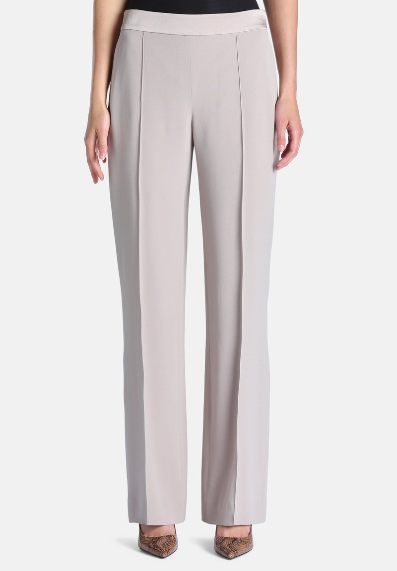 Femme portant un pantalon droit beige clair sur mesure avec des plis devant et des talons pointus en cuir de serpent sur fond blanc.
