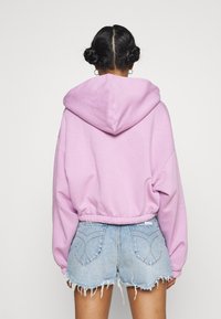 Hoodie rose court avec capuche, manches longues et taille froncée, associé à un short en denim déchiré. Texture lisse, design décontracté.