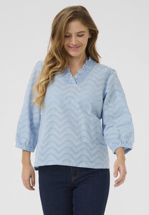 Blouse bleu clair avec un motif zigzag texturé, décolleté en V froncé et manches bouffantes. Le matériau semble doux et léger.