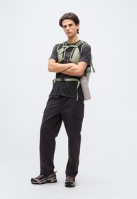 Νέος άνδρας όρθιος με σταυρωμένα τα χέρια, φοράει μαύρο γραφικό μπλουζάκι, πράσινο γιλέκο πεζοπορίας με παγούρι, μαύρο παντελόνι cargo και παπούτσια πεζοπορίας.