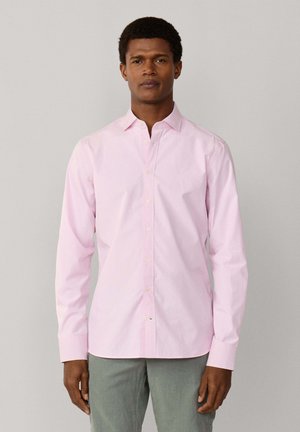 Hombre con camisa de manga larga de color rosa claro y pantalones grises, de pie frente a un fondo liso gris claro.