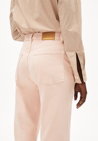 Jeans rose clair avec une texture lisse, dotés de poches arrière et d'une étiquette en cuir beige sur laquelle est inscrit "ARMEDANGELS". Chemise rayée visible.