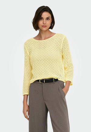 Femme portant un pull en maille crochet jaune clair à manches trois-quarts et un pantalon gris taille haute avec une ceinture noire.