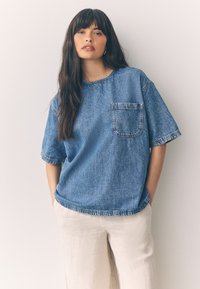 Maglietta oversize in denim blu con scollo rotondo e maniche corte, caratterizzata da una tasca frontale e orlo grezzo. Abbinata a pantaloni di colore chiaro.