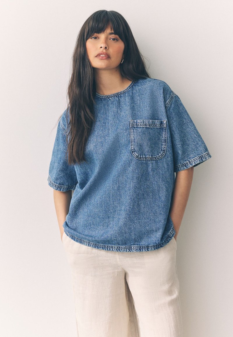 Blue denim oversized t-shirt with a round neckline and short sleeves, featuring a front pocket and raw hem detail. Paired with light-colored pants.